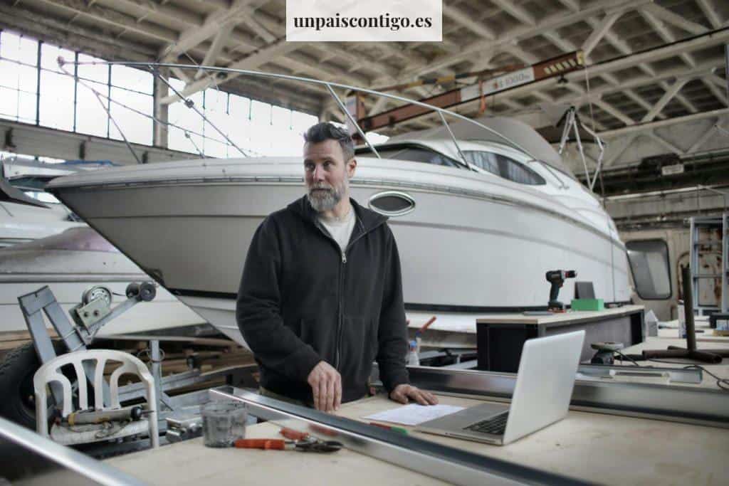 Adult bearded man in casual clothes using laptop while working with metal details near boat in workshop and looking away