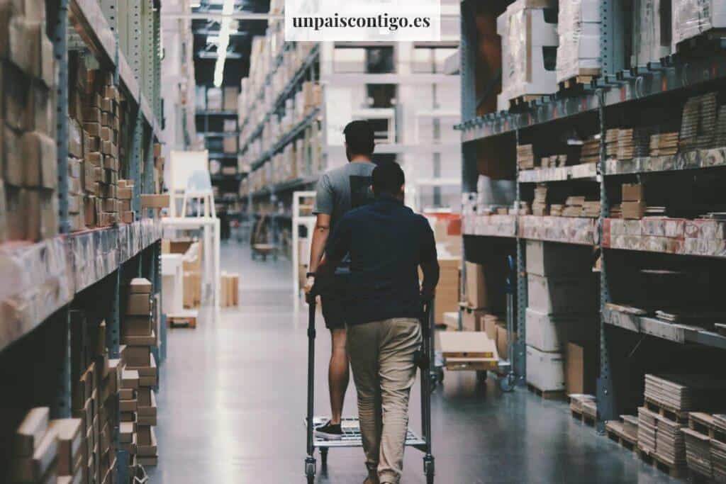 Two men maneuver a trolley in a large warehouse filled with boxes and shelves.
